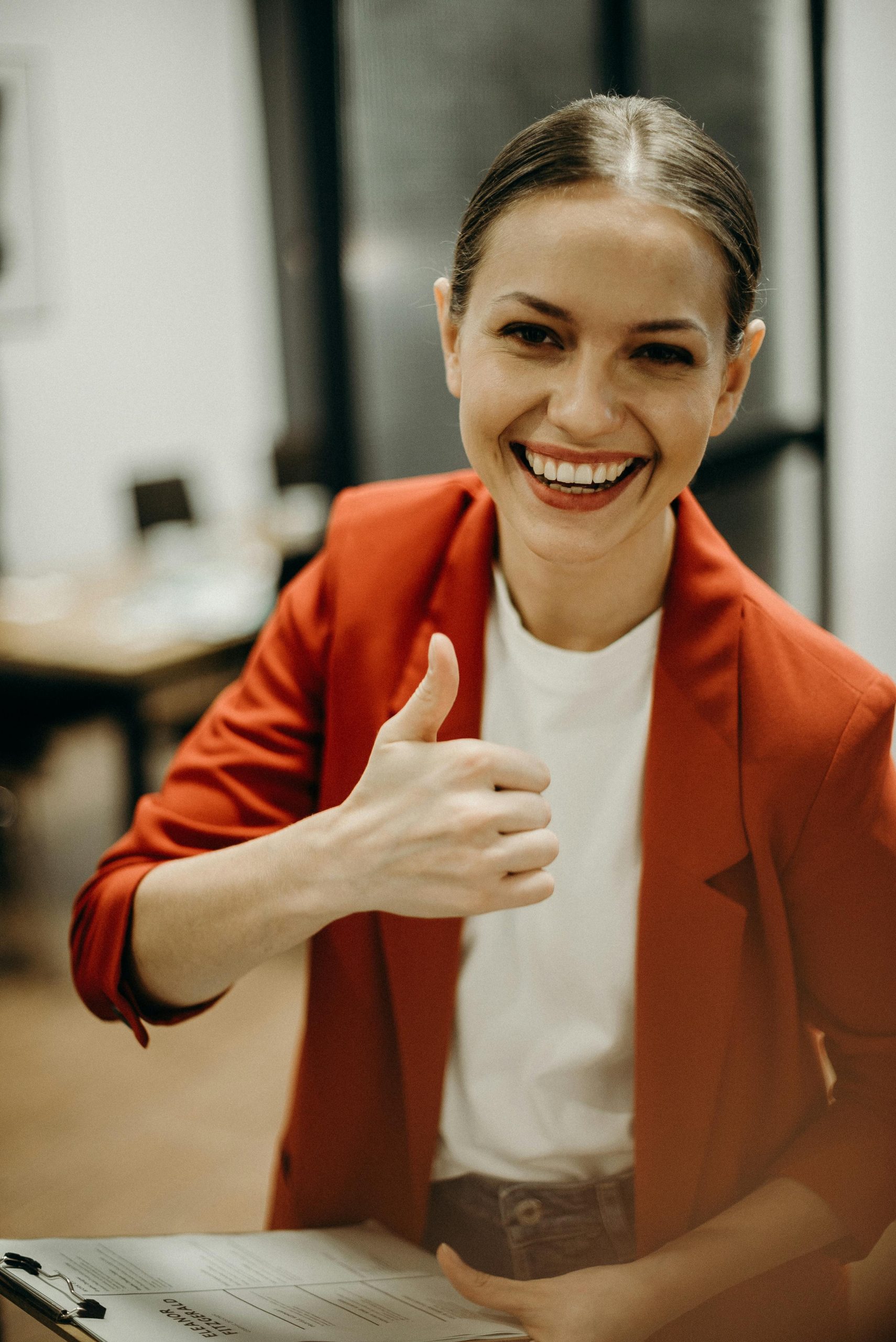 About Confident businesswoman in a red blazer gives a thumbs up in a modern office setting.